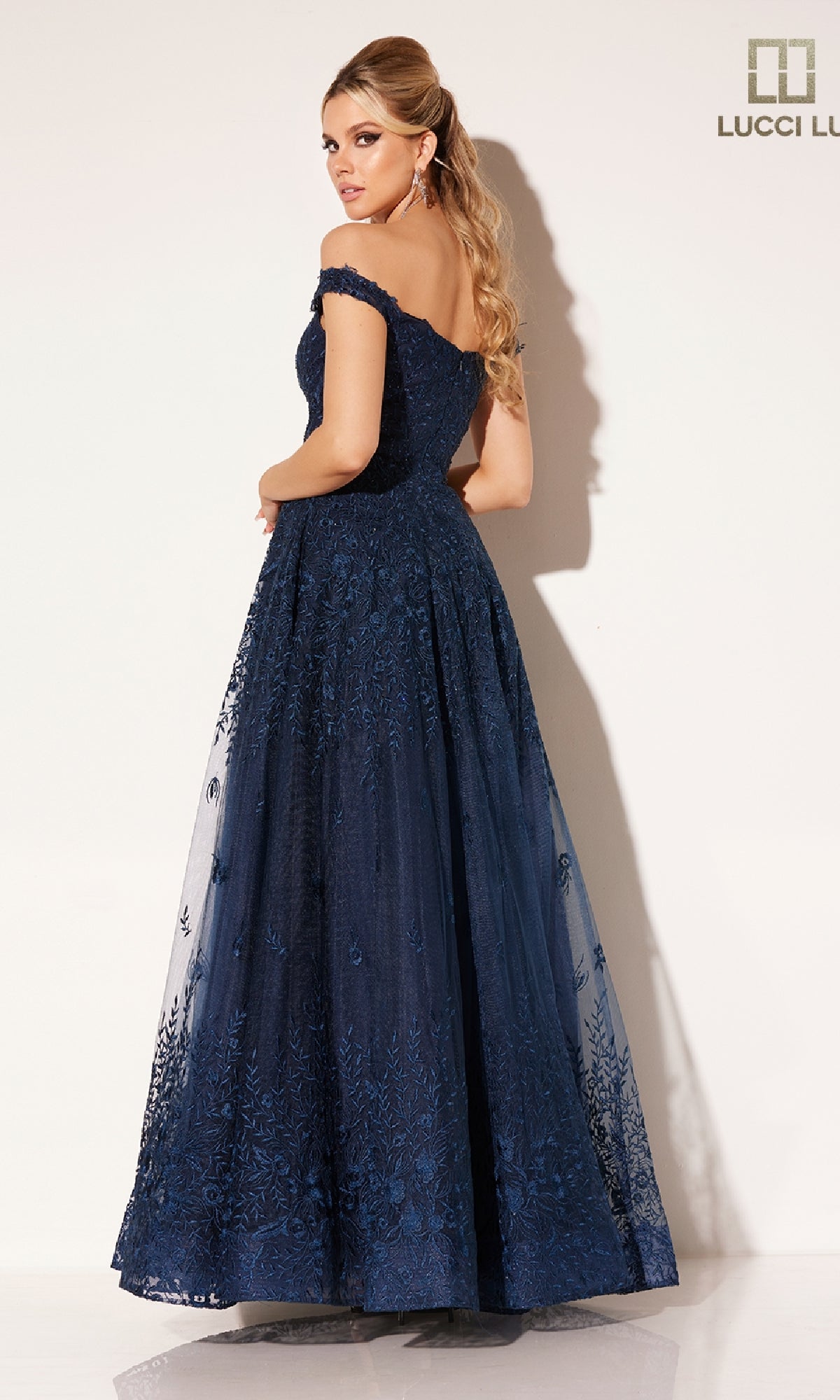 A woman poses against a plain light background, looking over her shoulder in the floor-length, navy blue Lucci Lu C8090 prom dress with off-the-shoulder lace detailing.